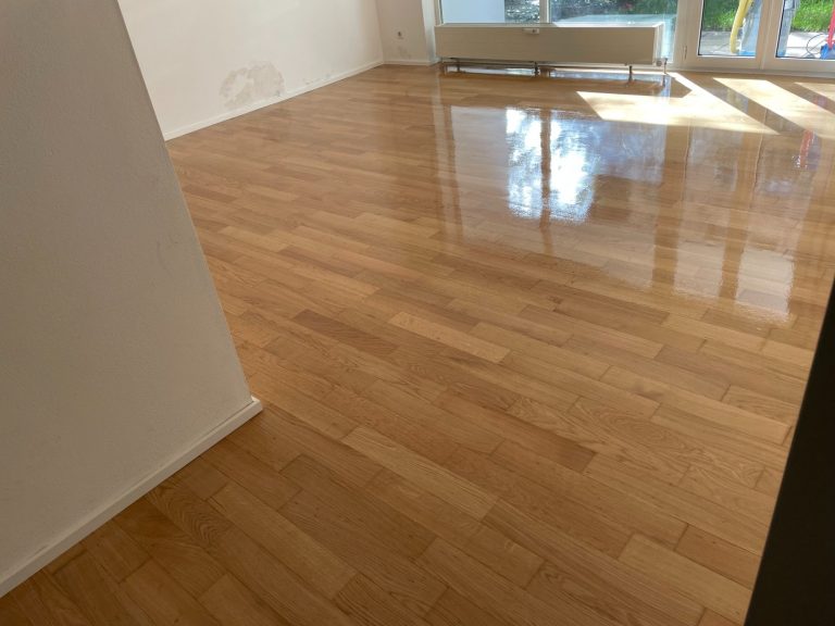 Heller Raum mit glänzendem, hellbraunem Parkettboden und großen Fenstern.