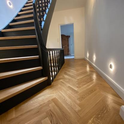 Holztreppe mit schwarzem Geländer, hölzernen Böden und sanfter Beleuchtung im Flur.